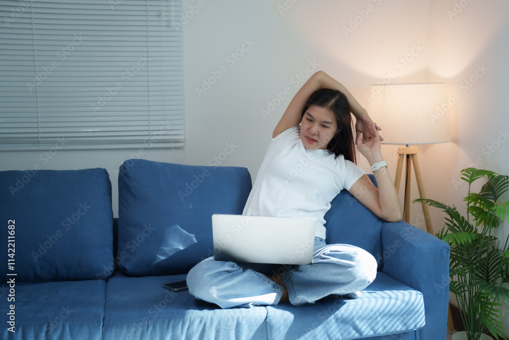 Young Asian woman is sitting on comfortable sofa and stretching her arms while working on laptop at home, taking a break from work to relax muscles Young Asian woman is sitting on comfortable sofa and stretching her arms while working on laptop at home, taking a break from work to relax muscles