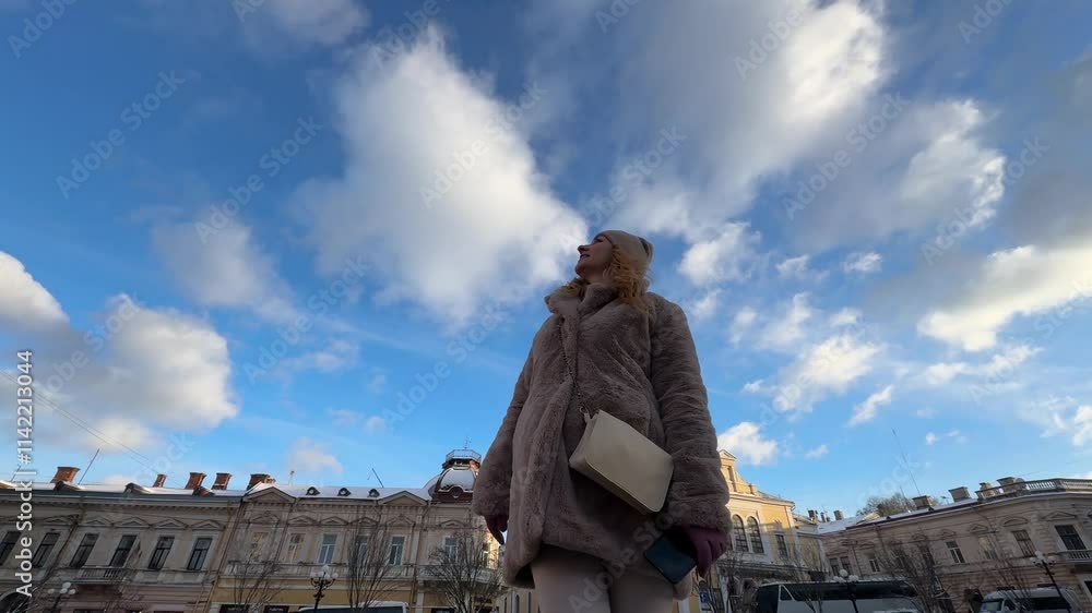 Woman in a fur coat poses happily in a city square under beautiful clouds and a bright blue sky Woman in a fur coat poses happily in a city square under beautiful clouds and a bright blue sky