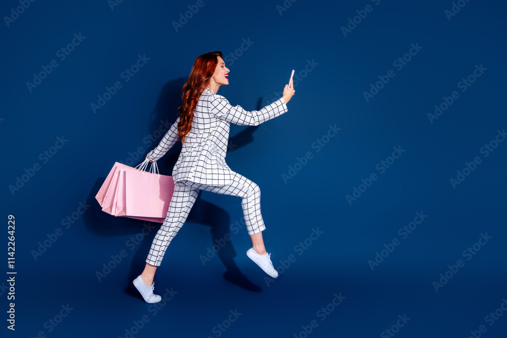 Confident young woman with red hair and checkered suit joyfully walking against dark blue background, holding shopping bags Confident young woman with red hair and checkered suit joyfully walking against dark blue background, holding shopping bags