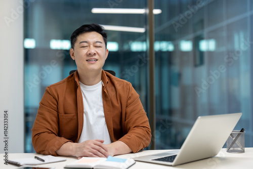 Asian male businessman meditates with eyes closed, promoting relaxation and mindfulness at work.