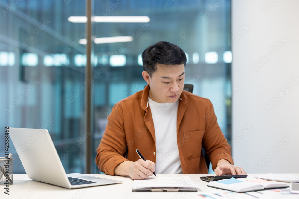 Asian man focused on completing tasks at his desk with a calculator and laptop, showcasing productivity in the workplace. Asian man focused on completing tasks at his desk with a calculator and laptop, showcasing productivity in the workplace.