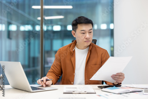 Asian man mature businessman in an office setting reviewing paperwork while working on a laptop, representing productivity, professionalism, and focus in a corporate environment.