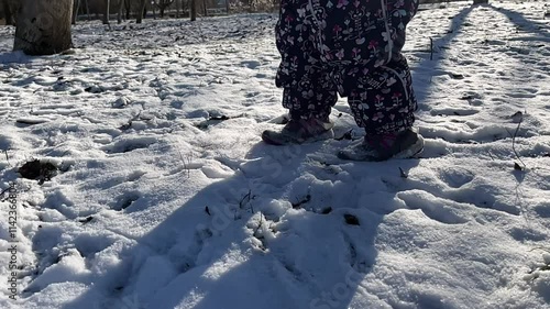 A small child walks through fresh snow. A girl in a down jacket steps on the snow and leaves footprints. Legs close up. Slow motion.