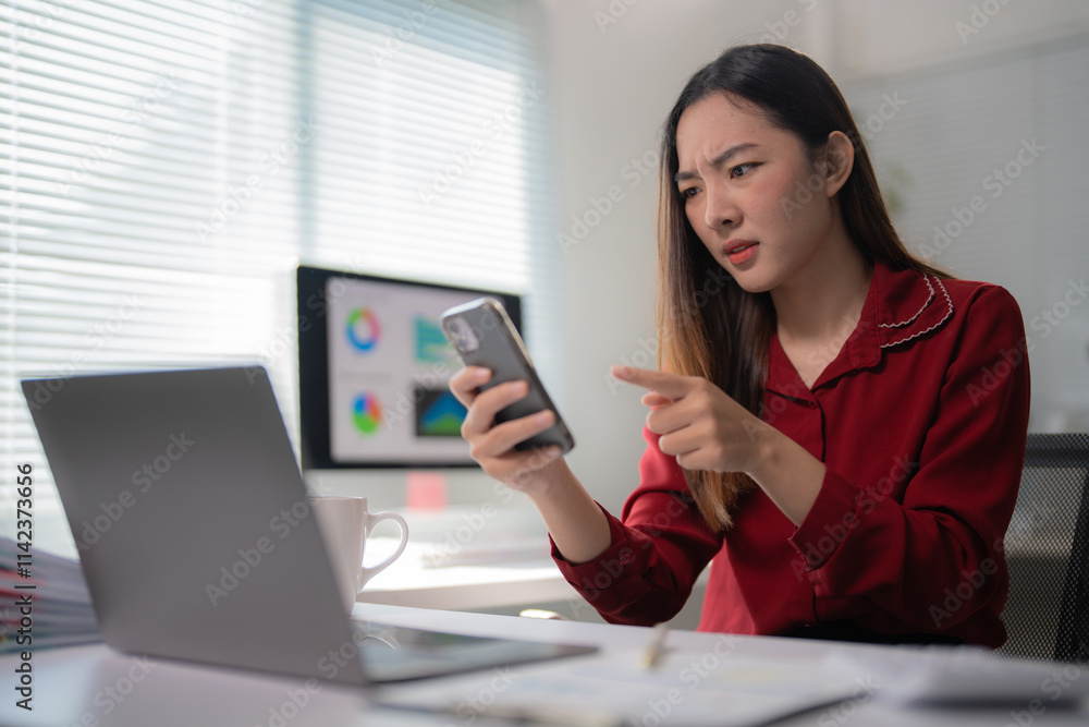 Young professional woman reading bad news on mobile phone while working with laptop and desktop computer at office, feeling stressed and confused by data on smartphone