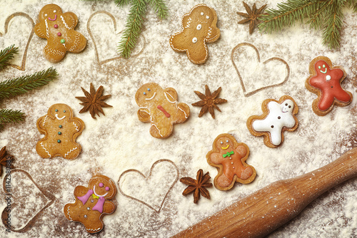Embellished with icing, a cheerful assortment of gingerbread cookies is set on a floured surface, surrounded by pine branches, star anise, and heart shapes, evoking a warm holiday spirit. 