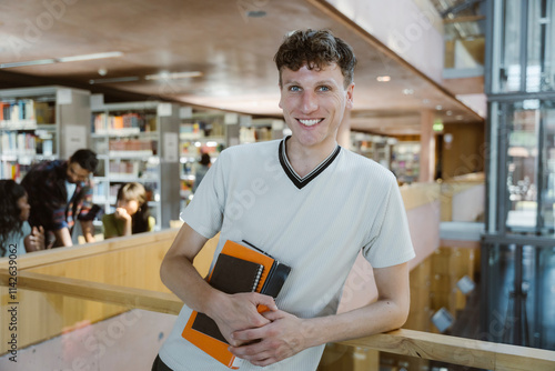 Portrait of smiling man holding books while leaning on railing in college library photo