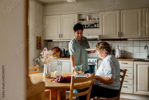 Smiling male caregiver serving breakfast to senior woman sitting at dining table