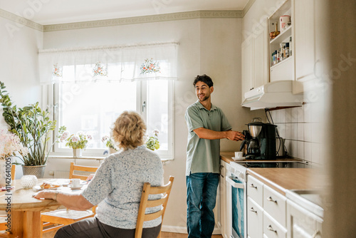 Male caregiver using coffee maker while talking with senior woman in kitchen at home photo