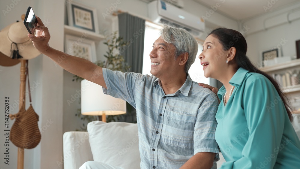 Fototapeta premium Happy grandfather and grandmother making video call with their son at home. Elder man holding smart phone and sitting at sofa while old woman greeting with cheerful vibe at modern house. Myrmidon.