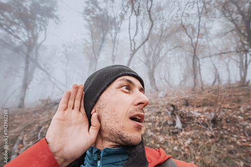 Male hiker cupping his hand to his ear, listening intently for sounds in a foggy forest, demonstrating alertness and caution photo