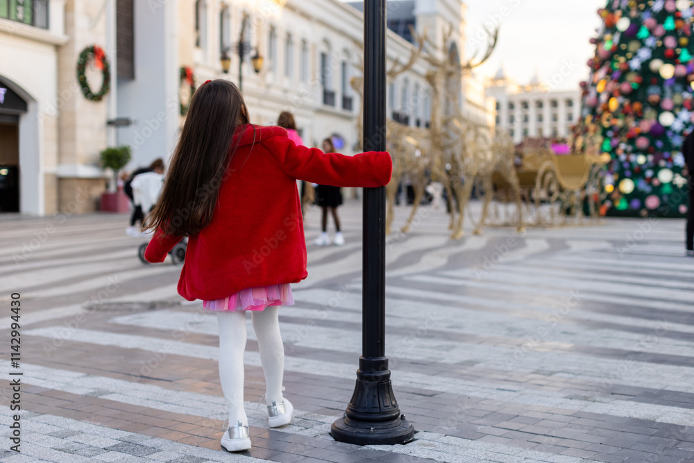 Naklejka premium A girl in a red coat twirls around a lamppost in a Christmas square, surrounded by decorations and a giant Christmas tree.