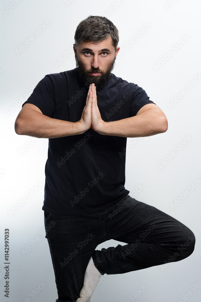 © Niko_Dali - A bearded man wearing a black shirt and dark jeans stands on one leg against a light background. He holds his hands together in a prayer position, maintaining balance. © Niko_Dali - A bearded man wearing a black shirt and dark jeans stands on one leg against a light background. He holds his hands together in a prayer position, maintaining balance.