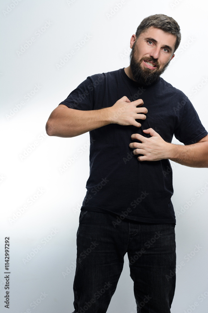 A man with a beard, wearing a black shirt and jeans, scratches his chest with both hands. He appears uncomfortable, suggesting skin irritation or allergy. A man with a beard, wearing a black shirt and jeans, scratches his chest with both hands. He appears uncomfortable, suggesting skin irritation or allergy.