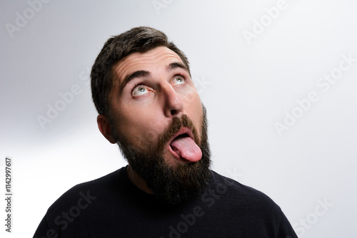 A bearded man looks upwards with his tongue sticking out against a plain background. He appears to be making a silly or playful expression.