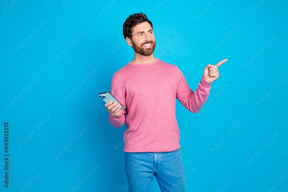 Confident bearded man in casual attire holding smartphone and pointing, standing against vibrant blue backdrop Confident bearded man in casual attire holding smartphone and pointing, standing against vibrant blue backdrop