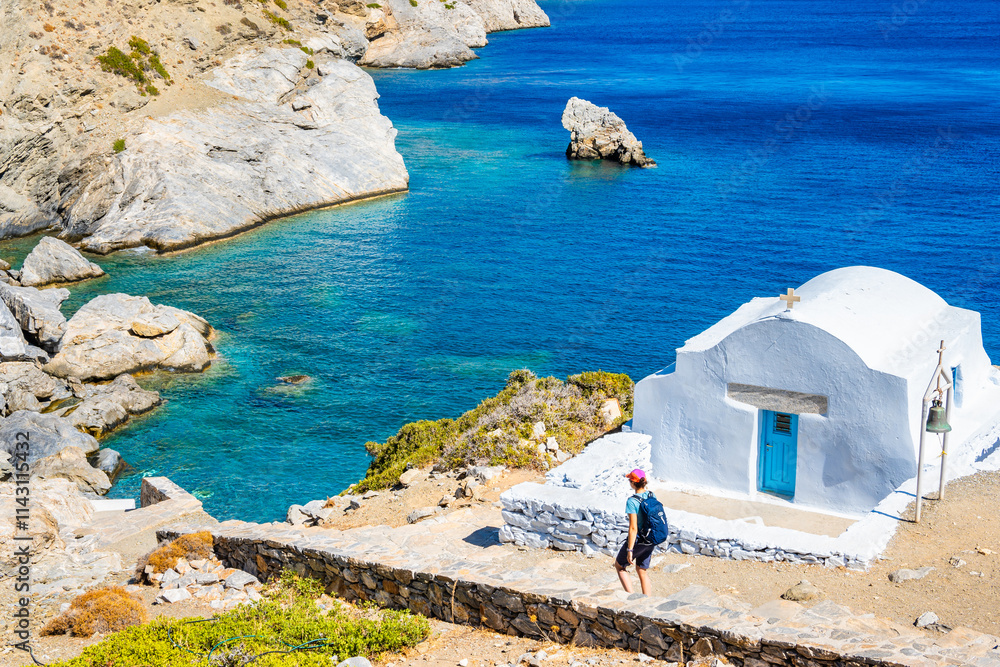 Woman tourist with backpack walking on path to Agia Anna beach and passing by white small Greek chapel, Amorgos island, Cyclades, Greece