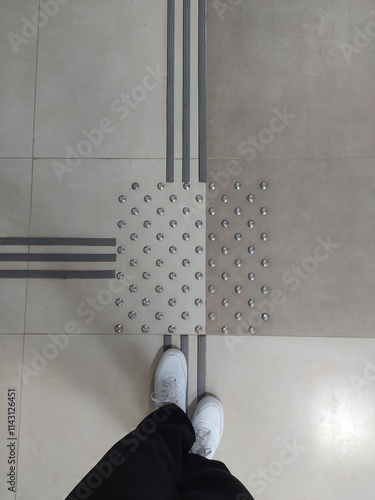 Feet in white sneakers in front of tactile tiles for visually impaired people on the floor indoors.