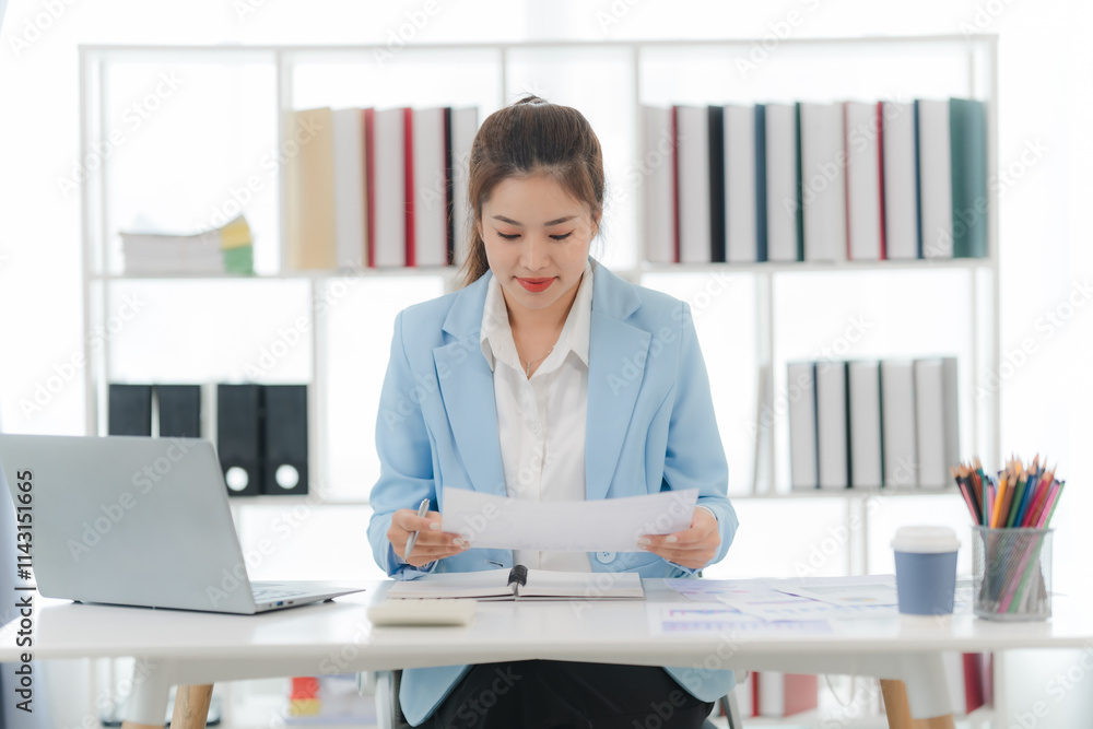 Focused and Confident: A young businesswoman in a sleek blue blazer meticulously reviews documents at her desk, demonstrating professionalism and determination in a modern office setting. Focused and Confident: A young businesswoman in a sleek blue blazer meticulously reviews documents at her desk, demonstrating professionalism and determination in a modern office setting.