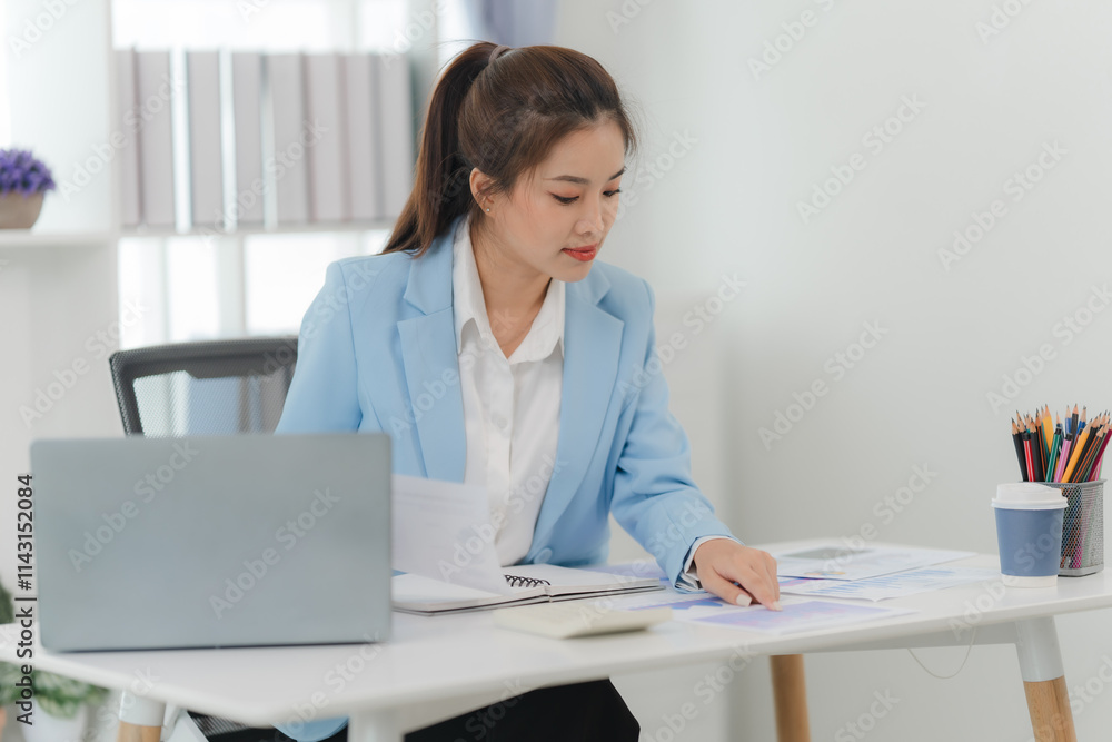 Focused Businesswoman Reviewing Documents: A young, serious businesswoman in a light blue blazer sits at her desk, meticulously reviewing documents and financial charts. She is engrossed in her work. Focused Businesswoman Reviewing Documents: A young, serious businesswoman in a light blue blazer sits at her desk, meticulously reviewing documents and financial charts. She is engrossed in her work.