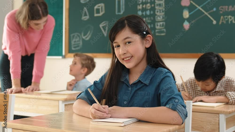 Asian girl writing test or classwork while looking at camera at classroom. Diverse happy student taking notes and teacher help caucasian boy doing test while sitting in front of blackboard. Pedagogy. Asian girl writing test or classwork while looking at camera at classroom. Diverse happy student taking notes and teacher help caucasian boy doing test while sitting in front of blackboard. Pedagogy.