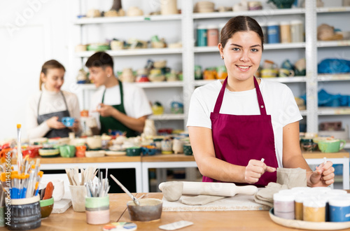 Interested young woman in apron cutting the edge of row clay cup with thread in pottery workshop photo