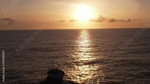 Imagem de um atuneiro navegando nas águas do Oceano Atlântico durante o pôr do sol, com o barco cortando suavemente as ondas sob a luz dourada e laranja do fim do dia. 