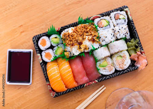 Against background of wooden table is rectangular plate of maki sushi set with uramaki and nigari rolls, decorated with imitation grass photo