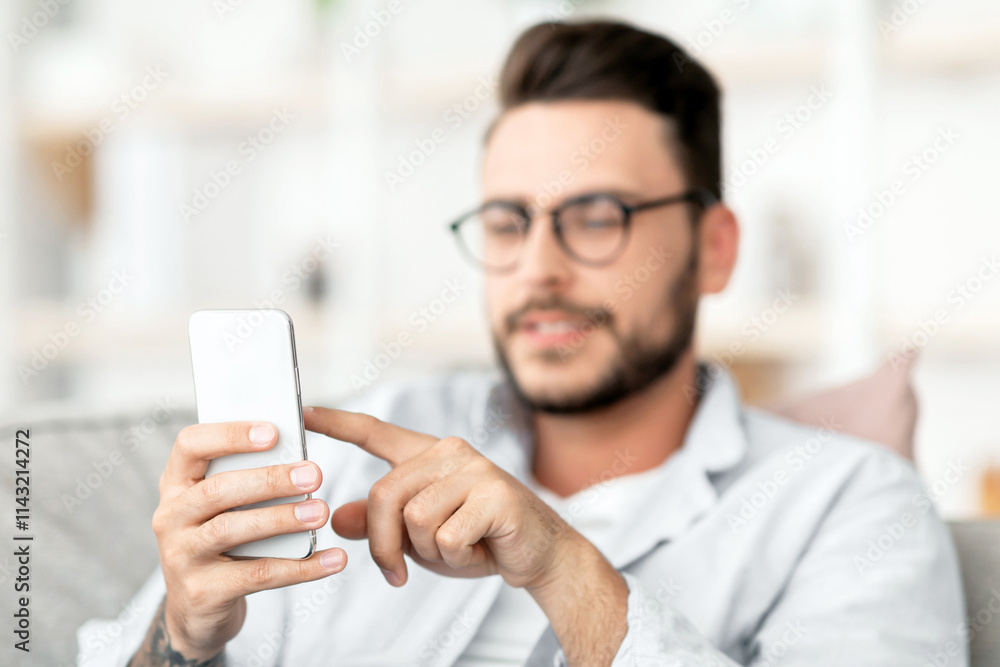 Handsome guy in eyeglasses using smatphone, touching screen while relaxing on couch at home, free space. Concentrated guy browsing internet or messaging with friends, sitting on sofa in living room Handsome guy in eyeglasses using smatphone, touching screen while relaxing on couch at home, free space. Concentrated guy browsing internet or messaging with friends, sitting on sofa in living room
