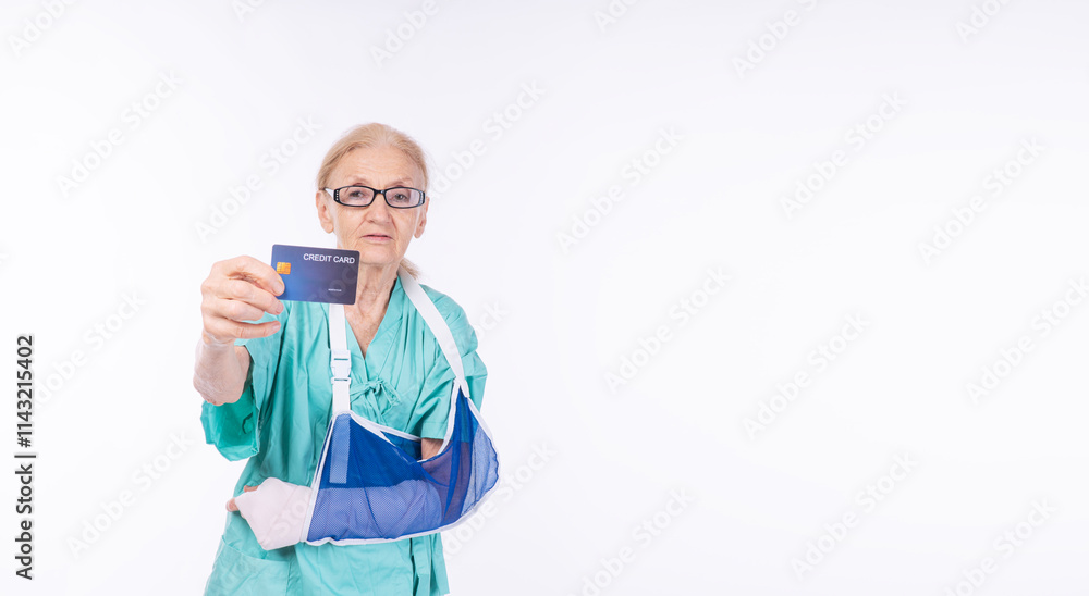 Injured grandmother wear glasses patent gown broken arm cast showing credit card payment standing over isolated white background. Elderly woman holding arm sling copy space. Insurance accident concept Injured grandmother wear glasses patent gown broken arm cast showing credit card payment standing over isolated white background. Elderly woman holding arm sling copy space. Insurance accident concept