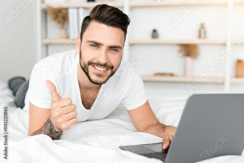 I like distant job. Millennial man with laptop computer gesturing thumbs up, working online, lying in bed at home. Freelance career and lifestyle, remote job concept