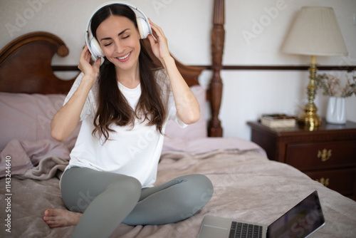 A young woman with long dark hair enjoys music through headphones while sitting on a cozy bed. She smiles, relaxed, dressed in casual attire, with a laptop nearby in a bright, stylish bedroom.