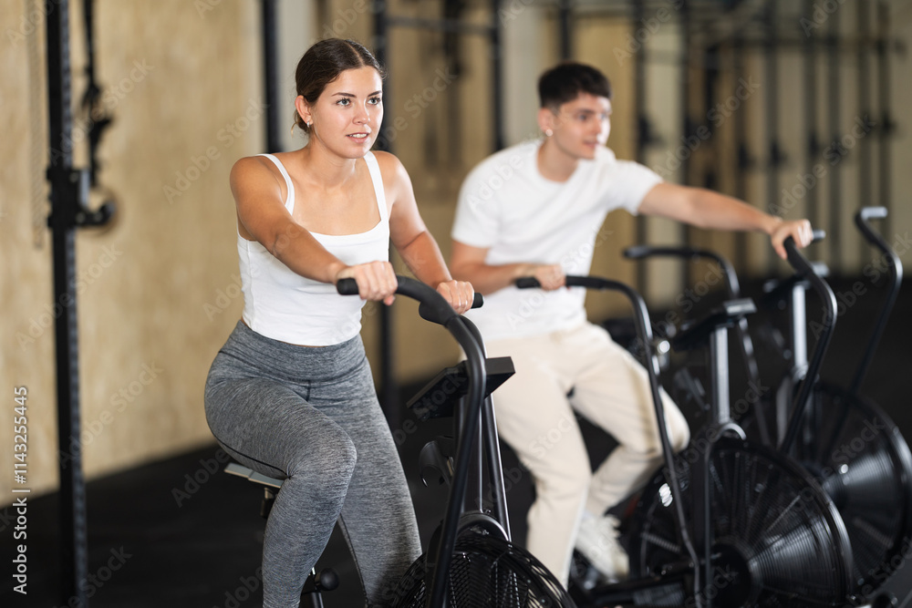 Positive girl riding exercise bike for cardio workout during cycling class at modern crossfit gym Positive girl riding exercise bike for cardio workout during cycling class at modern crossfit gym
