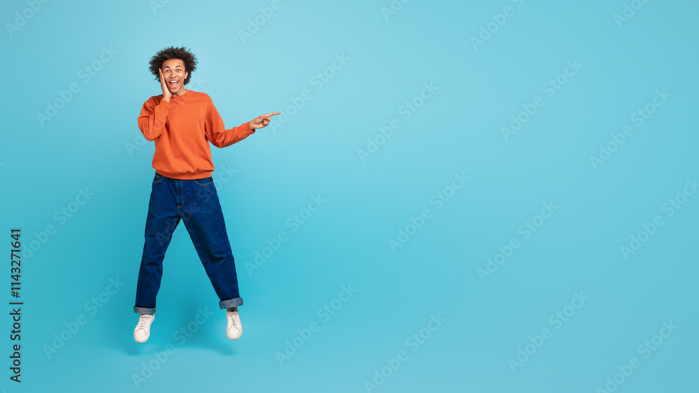 A young African American man with curly hair joyfully jumps in place, wearing a vibrant sweater and relaxed jeans, pointing at copy space A young African American man with curly hair joyfully jumps in place, wearing a vibrant sweater and relaxed jeans, pointing at copy space