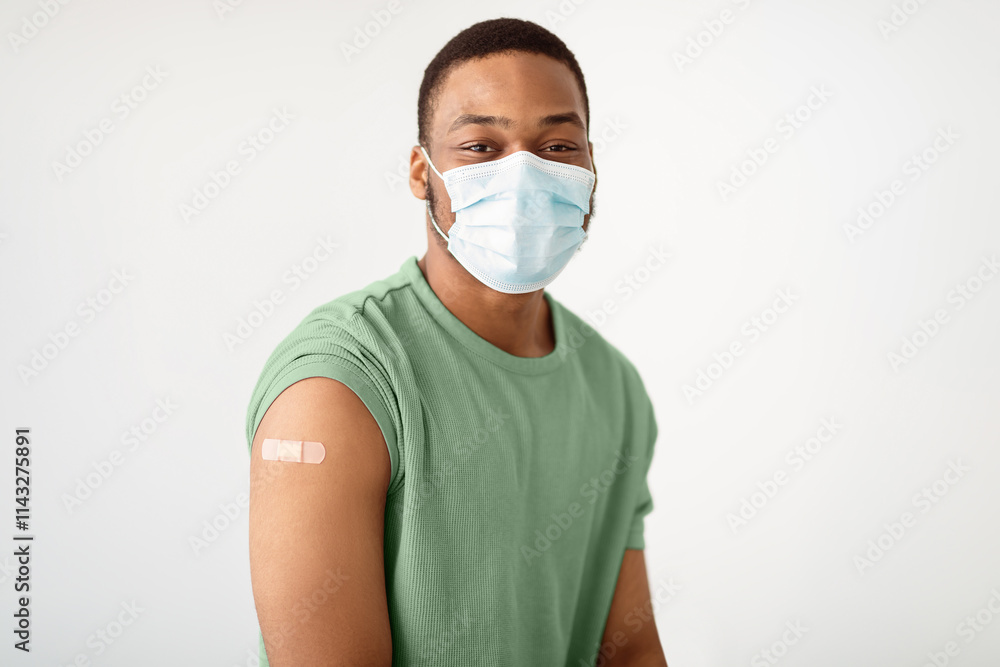 Coronavirus Vaccination. Portrait Of Vaccinated African Guy Wearing Protective Face Mask Looking At Camera Posing With Adhesive Tape On Arm After Vaccine Injection Over White Background Coronavirus Vaccination. Portrait Of Vaccinated African Guy Wearing Protective Face Mask Looking At Camera Posing With Adhesive Tape On Arm After Vaccine Injection Over White Background