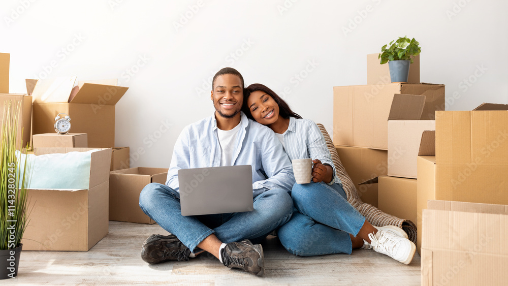 Moving concept. Happy african american couple purchasing furniture for new apartment, sitting on floor and using laptop computer, free space. Excited spouses smiling to camera, sitting among boxes Moving concept. Happy african american couple purchasing furniture for new apartment, sitting on floor and using laptop computer, free space. Excited spouses smiling to camera, sitting among boxes
