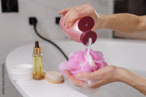 woman's hands applying shower gel on pink washcloth against bathroom background, body and hair care products lying nearby photo