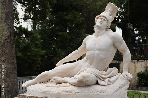 An antique statue of a reclining Achilles made of white stone with a layer of old dirt stands among the trees in a garden