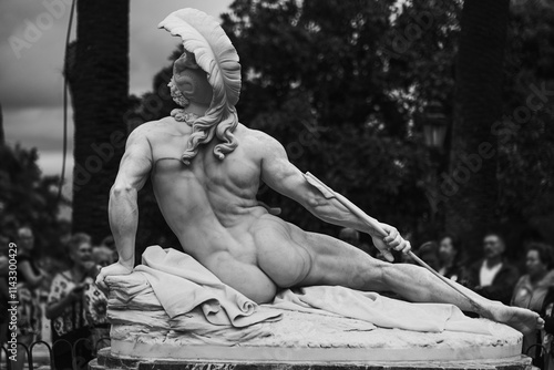 An antique statue of a reclining Achilles made of white stone with a layer of old dirt stands among the trees in a garden