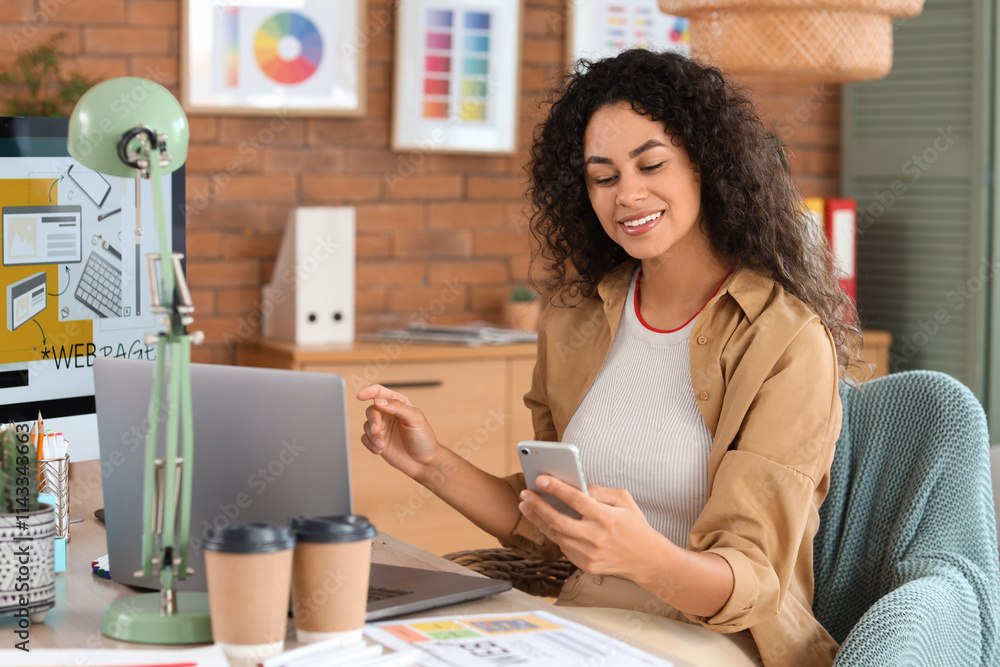 Female African-American web designer working with mobile phone at table in office Female African-American web designer working with mobile phone at table in office
