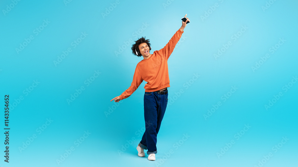 A young black man in a vibrant orange sweater playfully poses against a blue background. He holds a smartphone while expressing happiness and freedom, embodying a lively lifestyle. A young black man in a vibrant orange sweater playfully poses against a blue background. He holds a smartphone while expressing happiness and freedom, embodying a lively lifestyle.