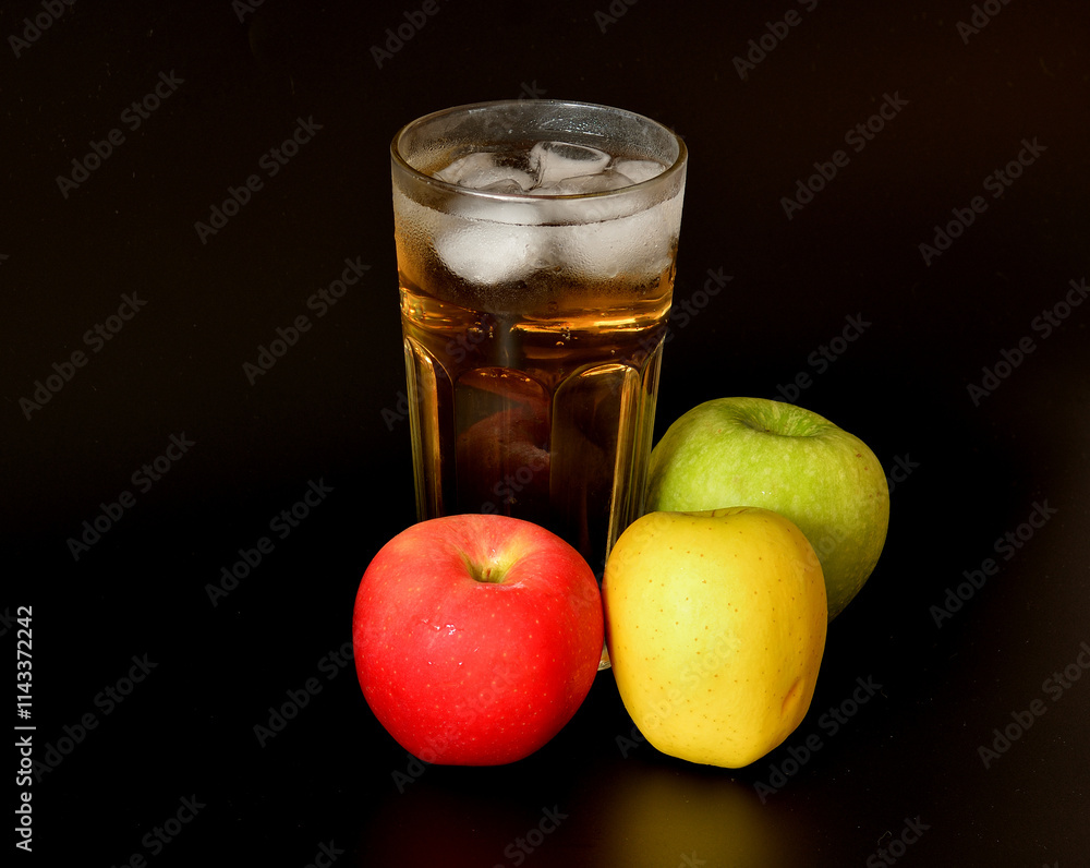 Apple juice in a tall faceted glass with ice on a black background, ripe fruits of different varieties nearby. Apple juice in a tall faceted glass with ice on a black background, ripe fruits of different varieties nearby.