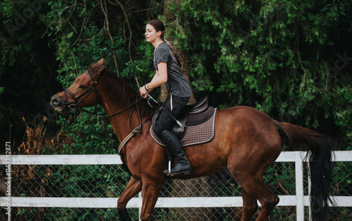 Young woman confidently riding a horse in an outdoor arena with lush green trees in the background. photo