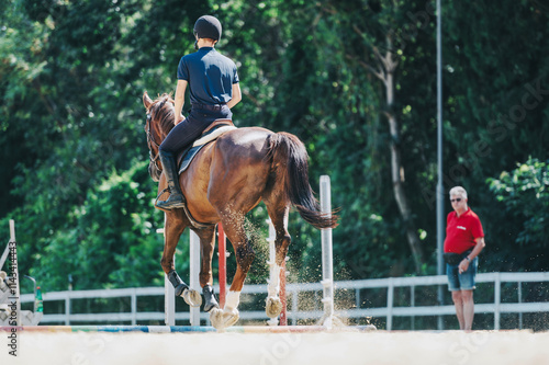 An equestrian rider in a helmet practices horseback riding in an outdoor arena. A coach observes from the background, highlighting focus and training in equestrian sports. photo