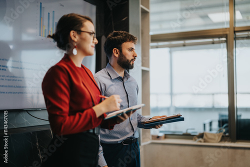 Two business people in a modern office environment discuss sales strategies, showcasing charts on a screen. The multicultural team collaborates with focus and engagement. photo