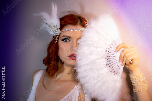 A woman with red hair holding a white fluffy scarf covering half of her face. Elegant retro clothing accessorized with a statement pearl bracelet and feather and sequin headdress.
 photo