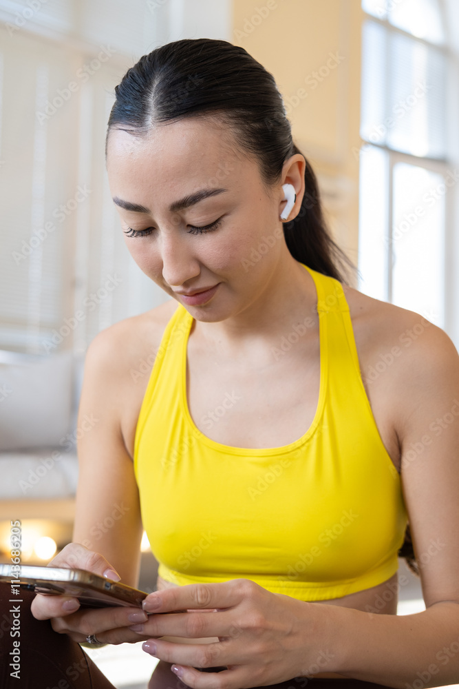 Happy athletic millennial asian woman in sportswear sitting on yoga mat in studio and smiling, holding smartphone or mobile phone, checking nice fitness mobile app. Listening to music in headphones Happy athletic millennial asian woman in sportswear sitting on yoga mat in studio and smiling, holding smartphone or mobile phone, checking nice fitness mobile app. Listening to music in headphones