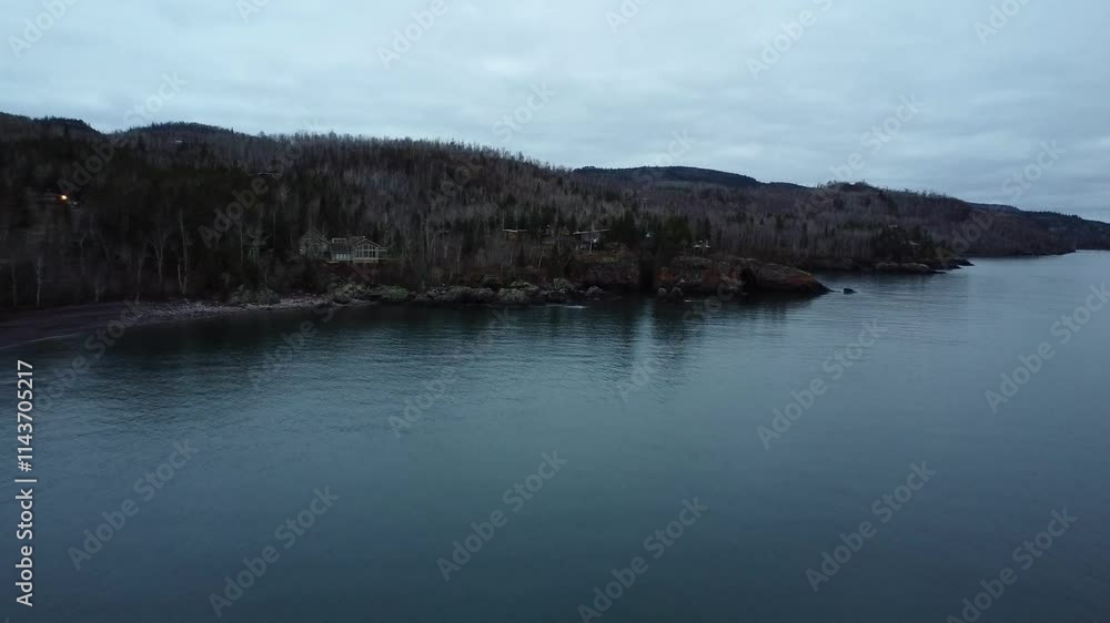 Serene cabain perched on a rocky lakeshore, surrounded by bare trees and evergreens, overlooking calm waters under a cloudy sky Serene cabain perched on a rocky lakeshore, surrounded by bare trees and evergreens, overlooking calm waters under a cloudy sky