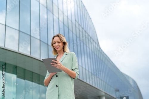 Confident White Businesswoman Using Tablet In Front a Modern Glass Office Building in the Business and Finance District, Smiling Happily while looking away