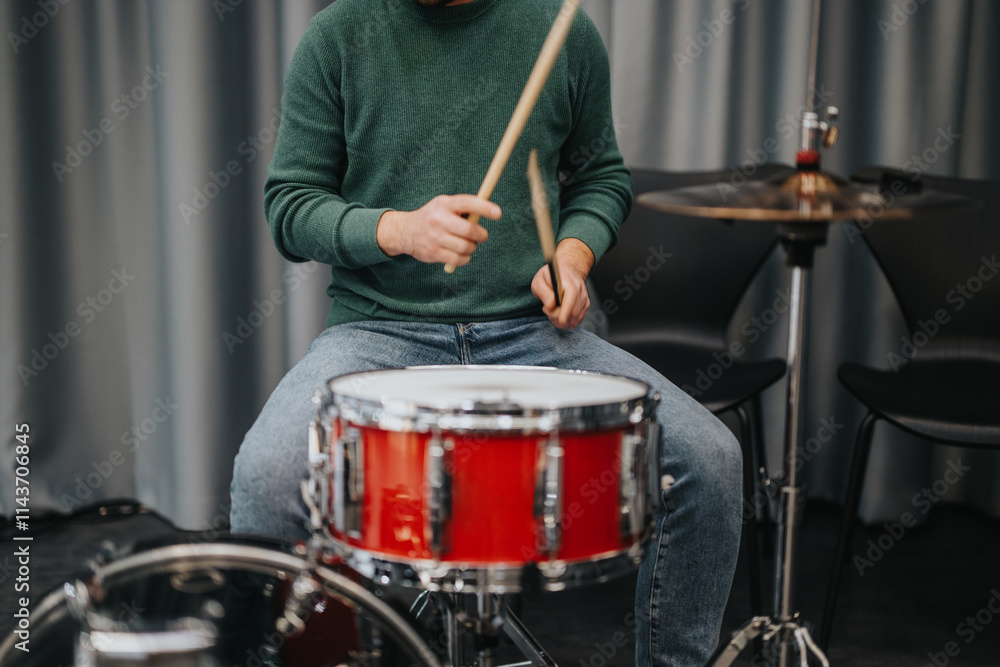 A musician wearing a green sweater is playing a red drum set. The focus is on the hands and drumsticks as they strike the drum in a studio environment. A musician wearing a green sweater is playing a red drum set. The focus is on the hands and drumsticks as they strike the drum in a studio environment.