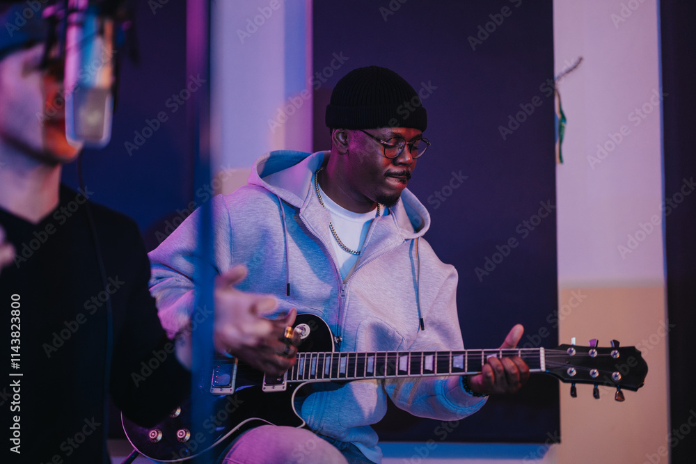 Obraz premium A young musician wearing a hoodie and beanie plays an electric guitar in a recording studio. The environment is professional, with a microphone visible in the foreground, highlighting music creation.